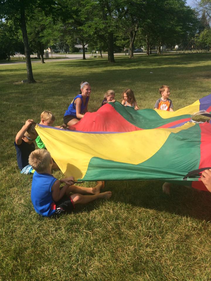 Youths and a parks counselor hold up a colorful tarp in a Village park